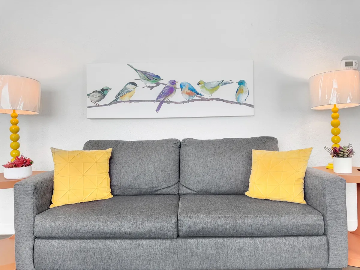 This inviting living area features a gray sofa accented with cheerful yellow pillows, bird-themed artwork, and stylish lamps. Large windows bring in natural light, creating a warm, social atmosphere perfect for group hangouts or game nights after a day out in the city.