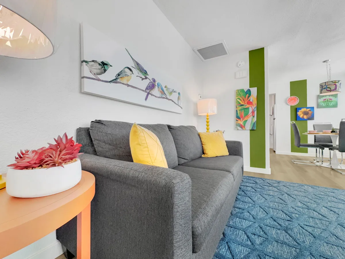 A cheerful living area featuring a gray sofa adorned with a yellow pillow, vibrant bird artwork on a green accent wall, and an orange side table. Soft daylight streams in, creating a welcoming spot for guests to relax after a day out exploring with their pet.