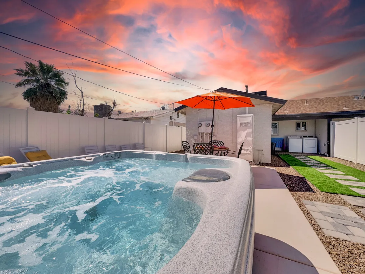 This image features a spacious private deck with a bubbling hot tub, an orange umbrella providing shade over a dining table, lush greenery, and a palm tree. The sunset sky adds a tranquil ambiance, and the setup invites both relaxing soaks and al fresco dining.