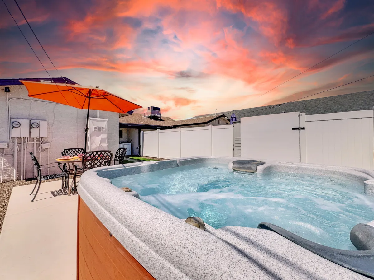 A welcoming deck area featuring a hot tub, comfortable outdoor seating, and a vibrant orange umbrella. The scene is lush with greenery and set at sunset, creating an inviting and tranquil mood ideal for relaxation or entertaining guests.
