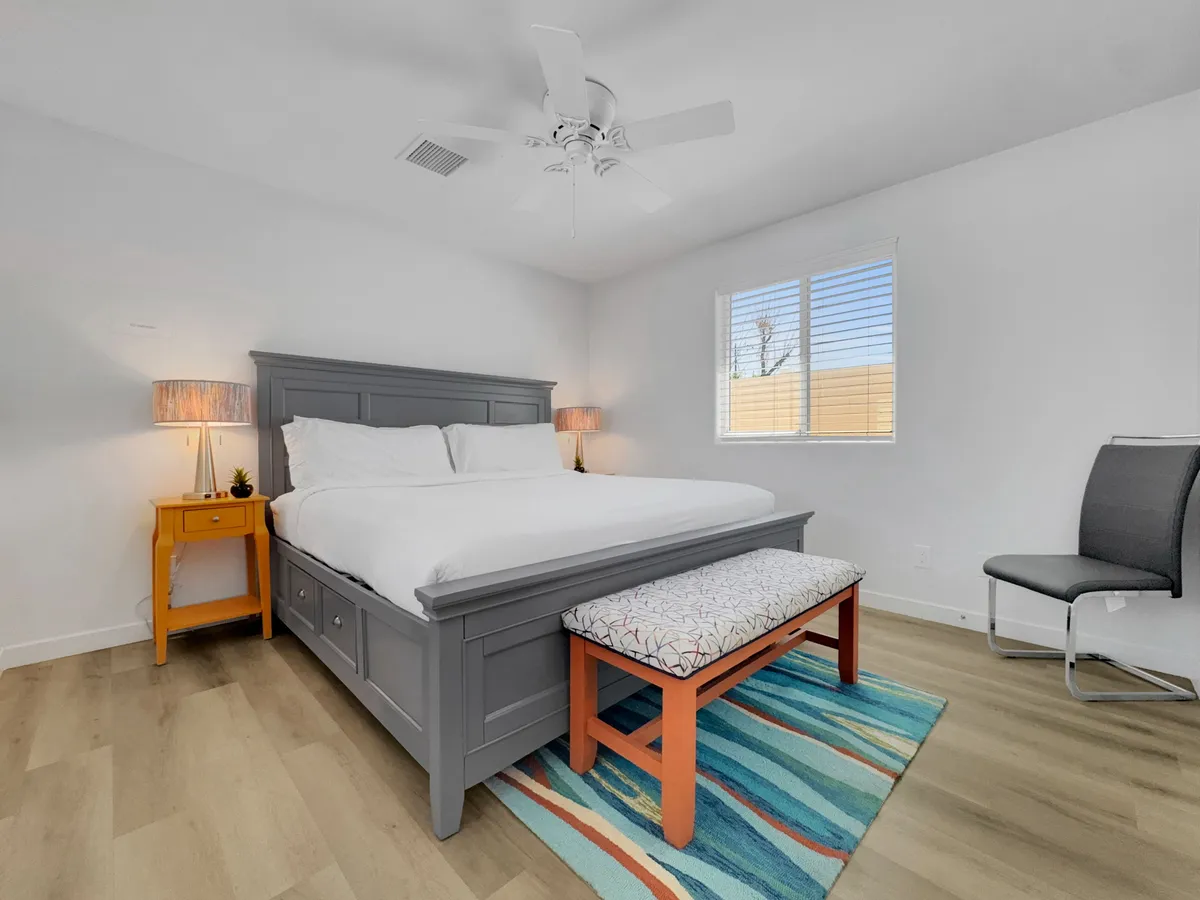 A tranquil bedroom featuring a neatly made bed with crisp linens, an orange nightstand, and a colorful rug that adds a touch of personality. A bench at the foot of the bed and soft natural light create a calm, peaceful mood ideal for relaxation.
