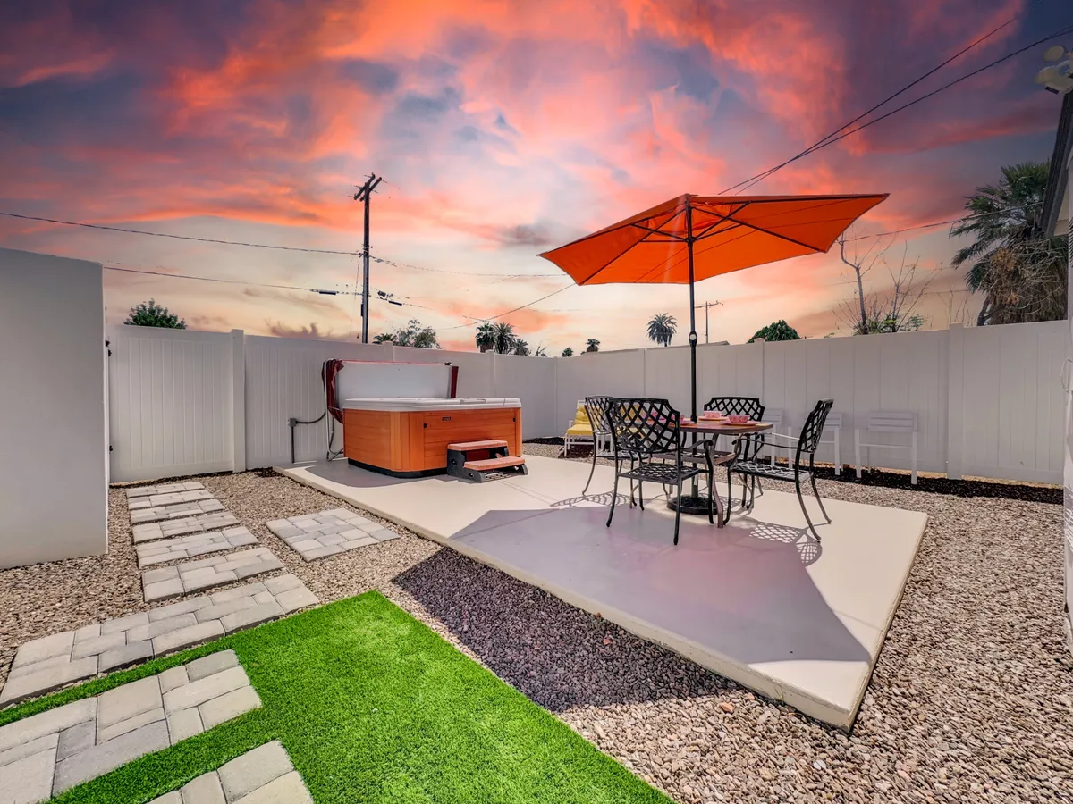 A serene deck scene with a hot tub, outdoor dining set beneath a parasol, and a winding pathway surrounded by greenery. The view suggests privacy and invites guests to unwind, dine, or socialize in a peaceful setting.