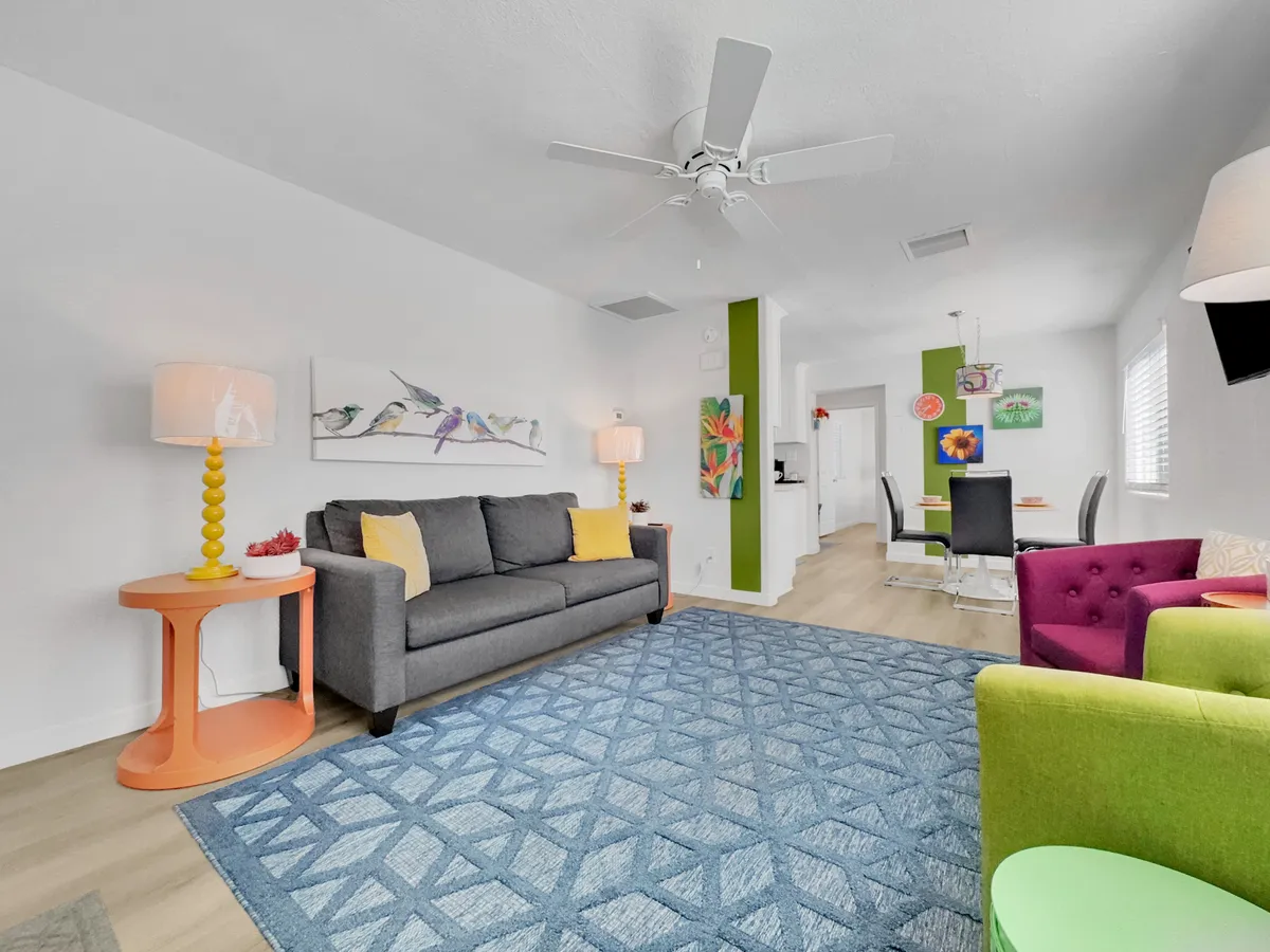 A plush gray sofa paired with playful, colorful accent chairs and an orange side table, all anchored by a bold blue area rug and spirited wall art. The arrangement invites relaxation and effortless conversation.