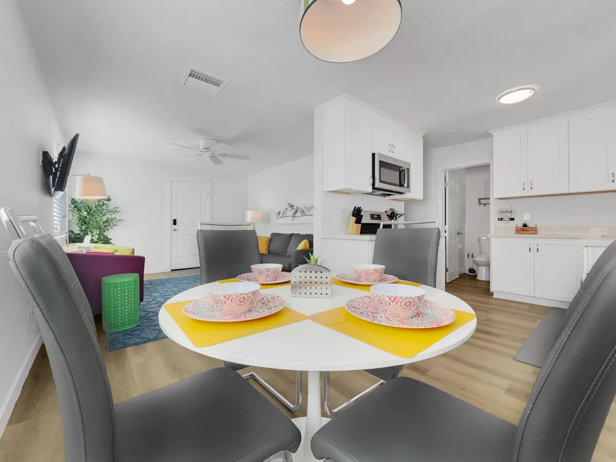 A welcoming open-plan kitchen and dining area, with a dining table set for a meal, colorful sofa, and kitchen in the background. The bright, cheerful atmosphere suggests a space where dining, socializing, and cooking blend effortlessly.